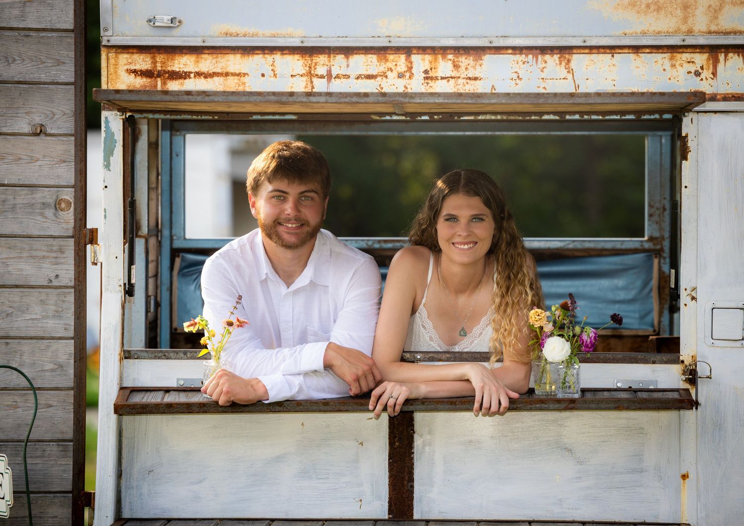 A smiling couple leans out of a rustic vintage trailer window adorned with wildflowers on a sunny day.