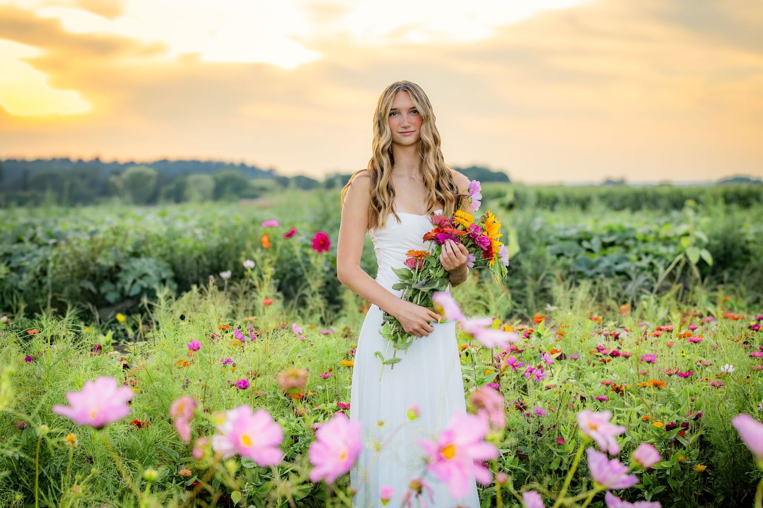 Young woman in white dress holding colorful bouquet stands in vibrant flower field at golden sunset.