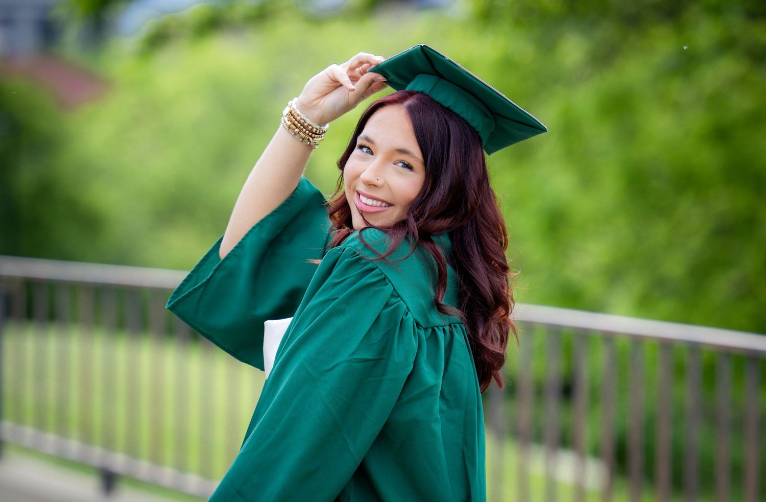 Smiling young woman in green graduation cap and gown posing outdoors with greenery in the background.