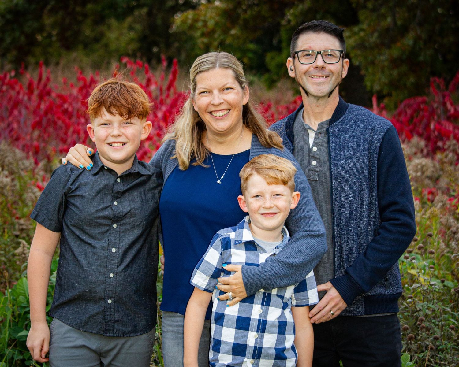 Family of four smiling together outdoors in front of vibrant red garden plants.