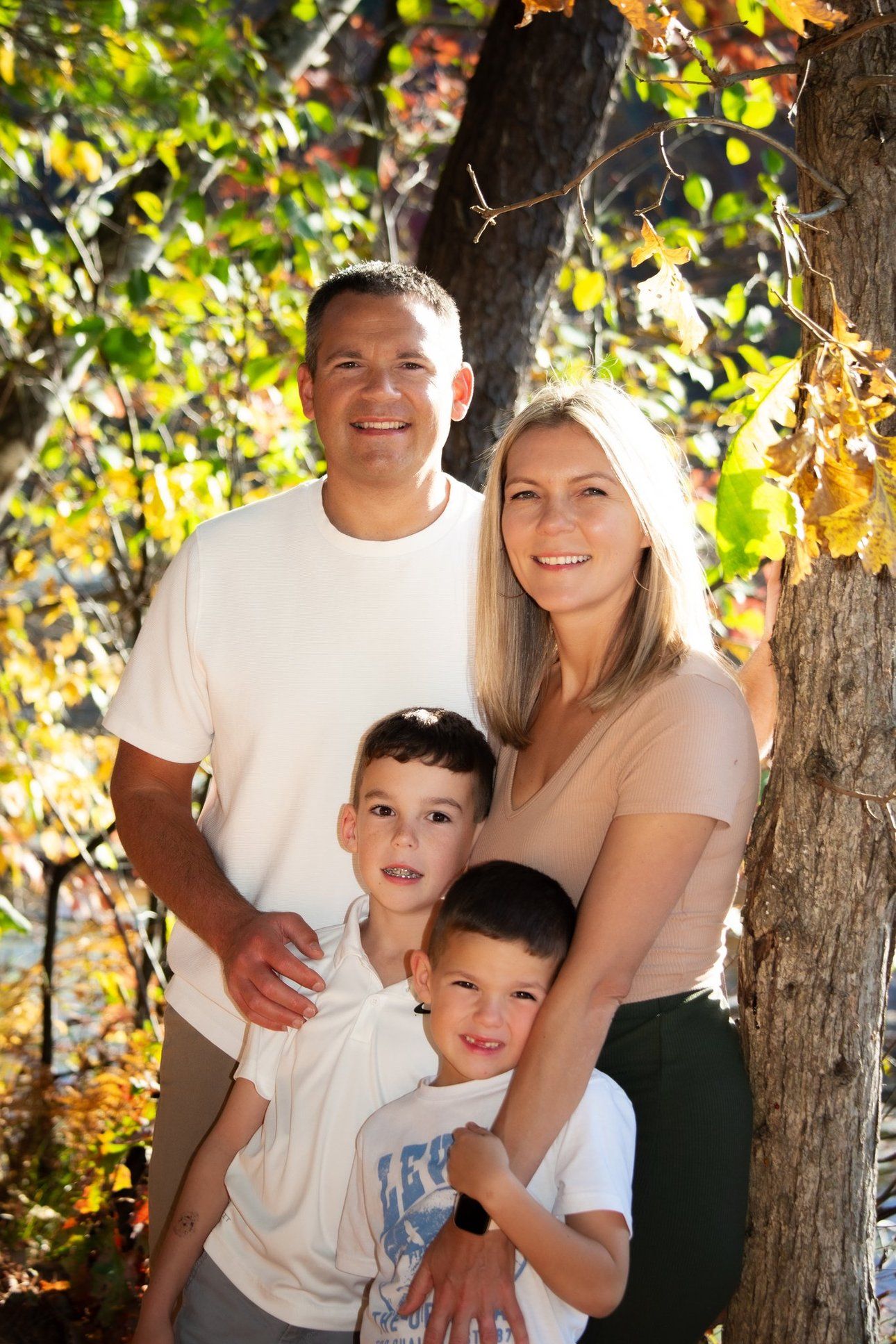 Happy family of four posing outdoors among colorful autumn foliage trees during fall season portrait session.
