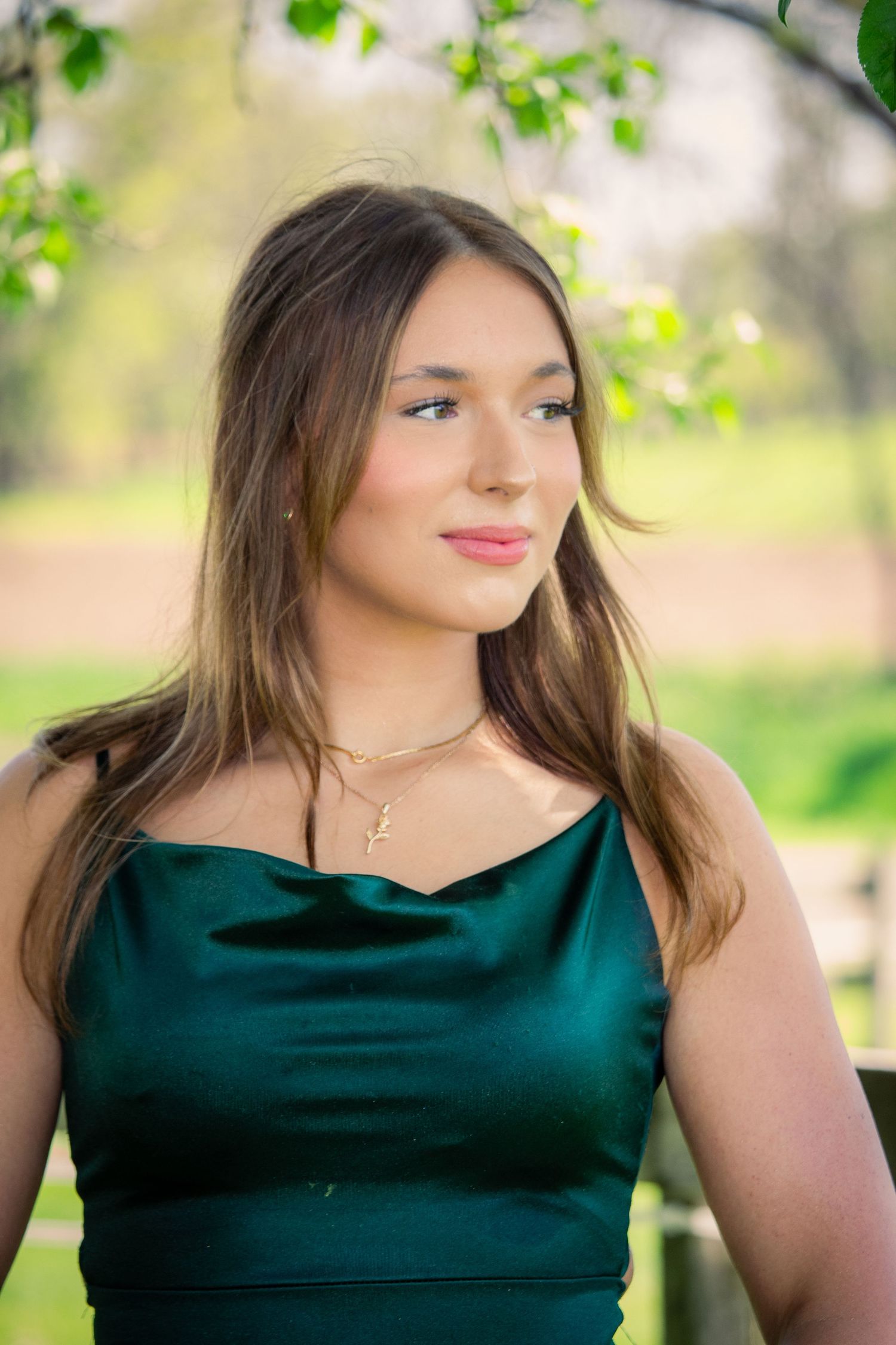 Beautiful young woman in emerald green satin dress posing outdoors under blossoming spring tree branches.