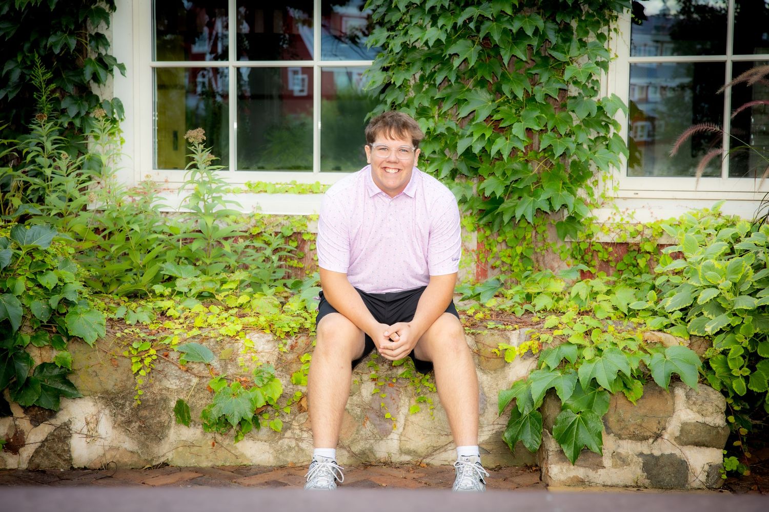 Young man in pink polo shirt and shorts sitting on a stone wall surrounded by lush green ivy outdoors.