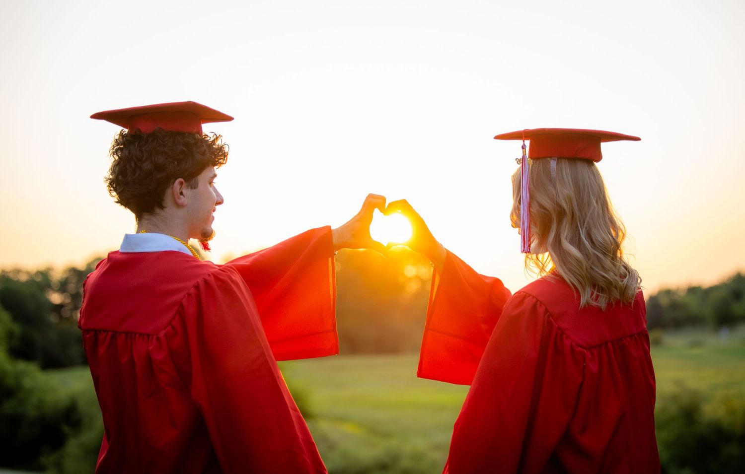 Two graduates in red robes making a heart shape with hands framing the golden sunset sky.