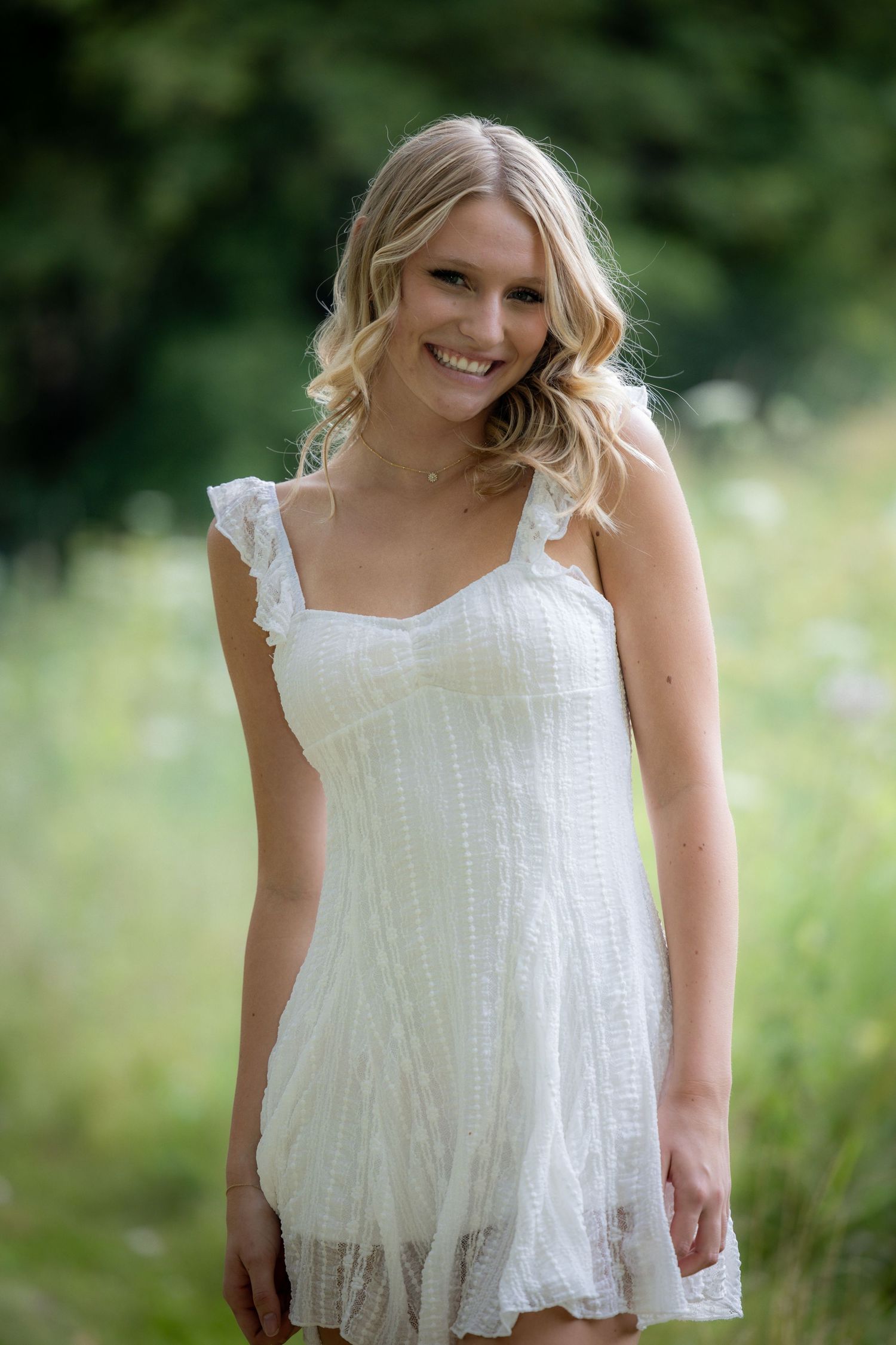 Cheerful blonde woman in white dress posing in a green outdoor setting.