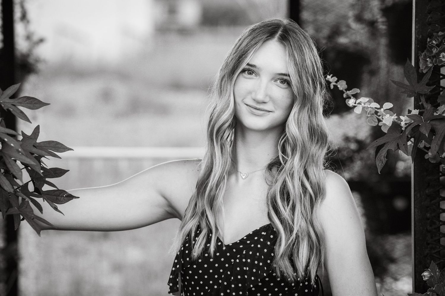 Young woman with long wavy hair smiling outdoors among leaves in a black and white portrait photo.