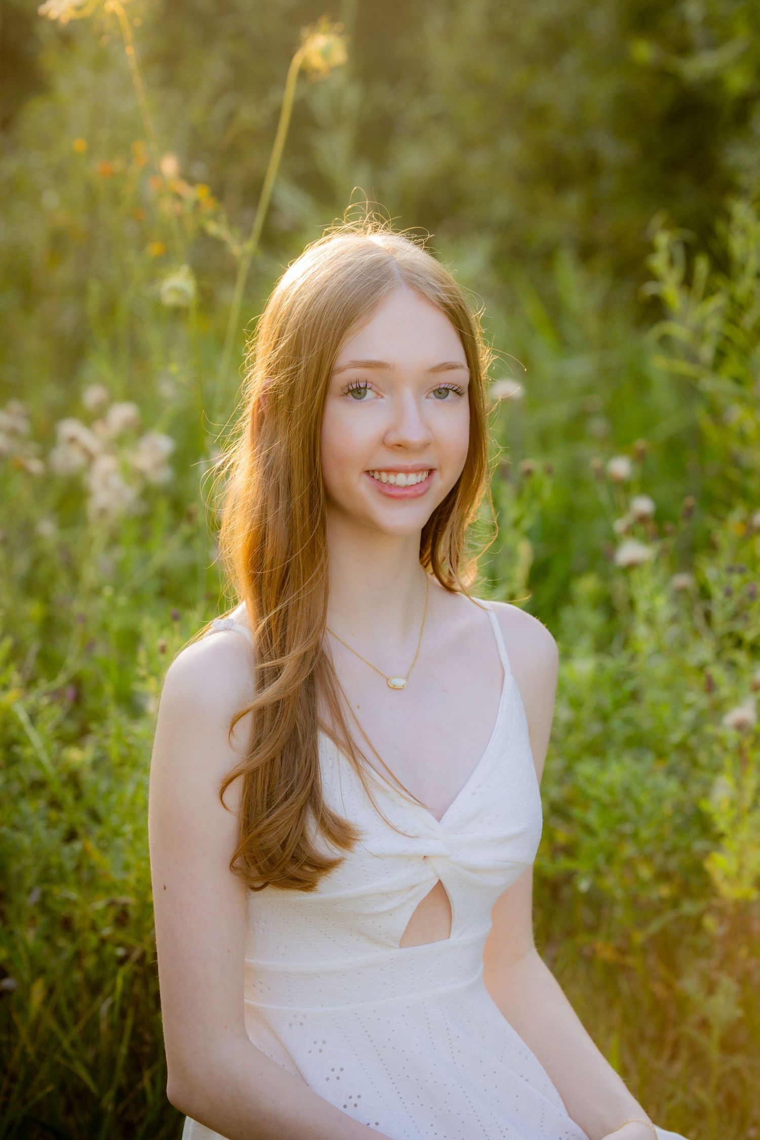 Young woman with long auburn hair smiling in a white sundress, surrounded by wildflowers in golden sunlight.