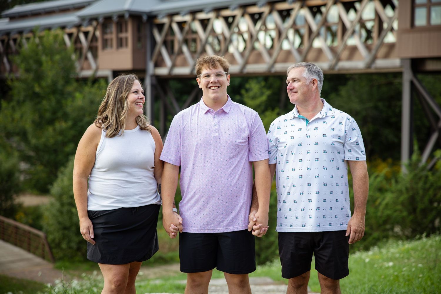 A smiling family of three poses outdoors near a wooden bridge surrounded by lush greenery.