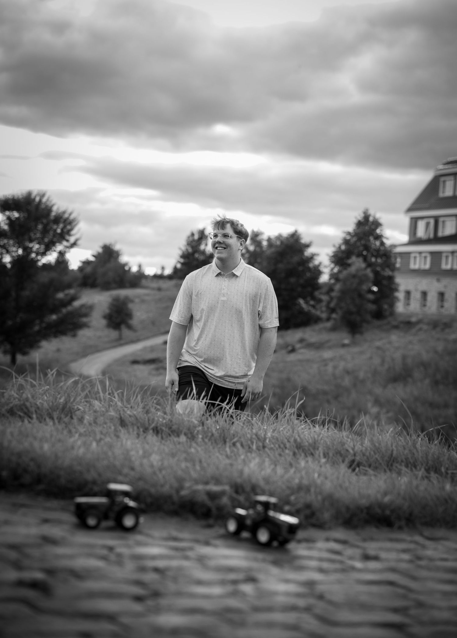 Black and white photo of a smiling man standing on a grassy hill with toy tractors in the foreground.