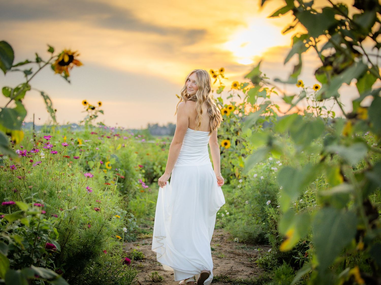 A blonde woman in a white dress walks through a sunflower field at golden sunset.