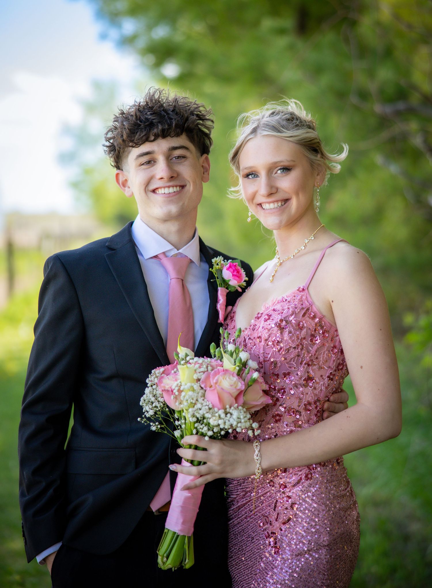 Smiling teenage prom couple poses outdoors, girl in pink sequin dress holding floral bouquet.
