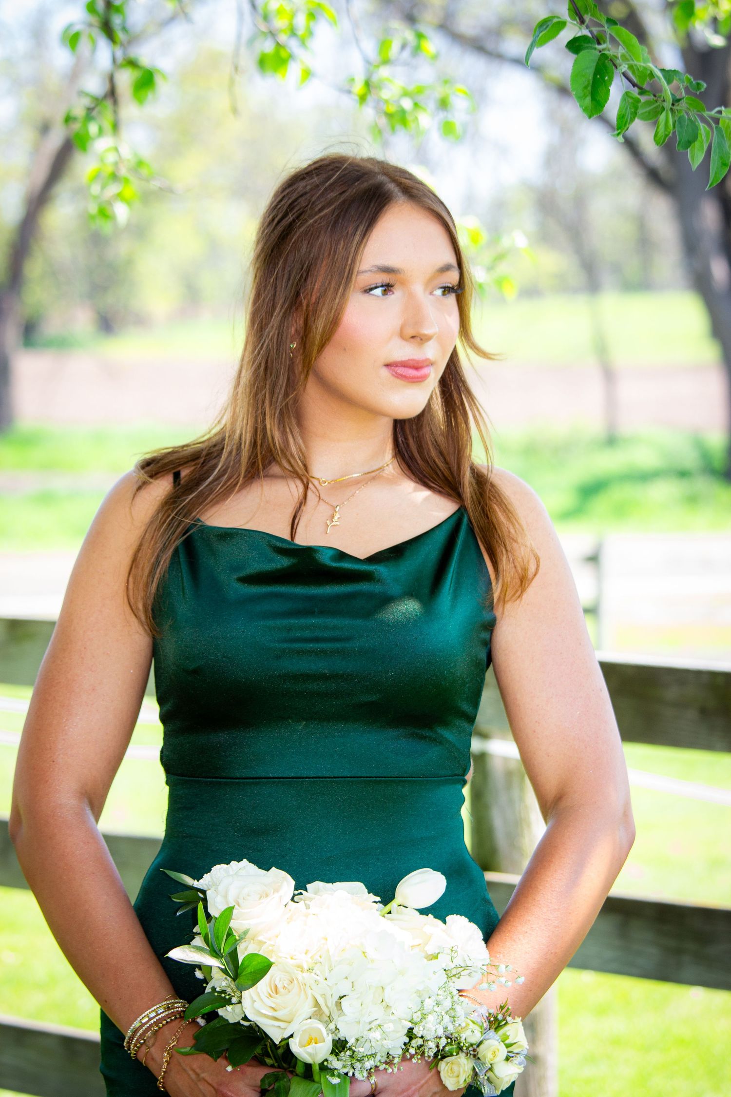 Young woman in emerald green gown holding white bouquet standing by wooden fence in garden setting.