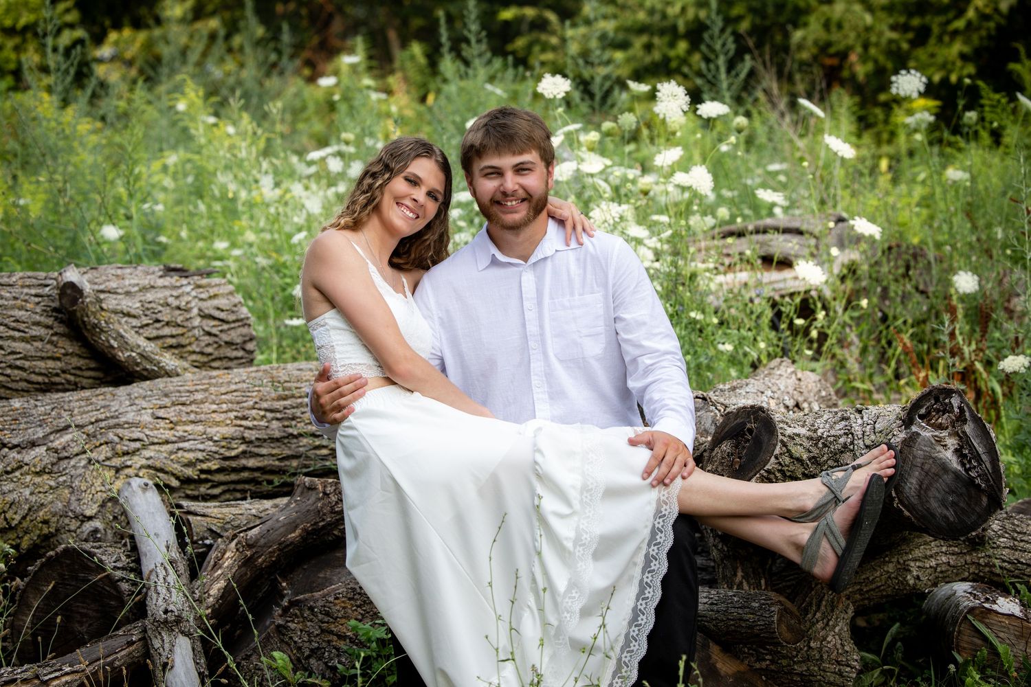 Happy couple in white attire sitting on logs surrounded by wildflowers in a lush garden setting.