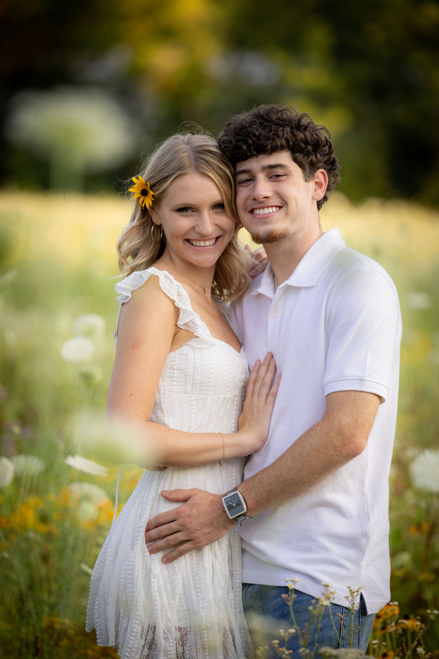A smiling young couple embraces in a vibrant wildflower meadow with golden sunlight behind them.