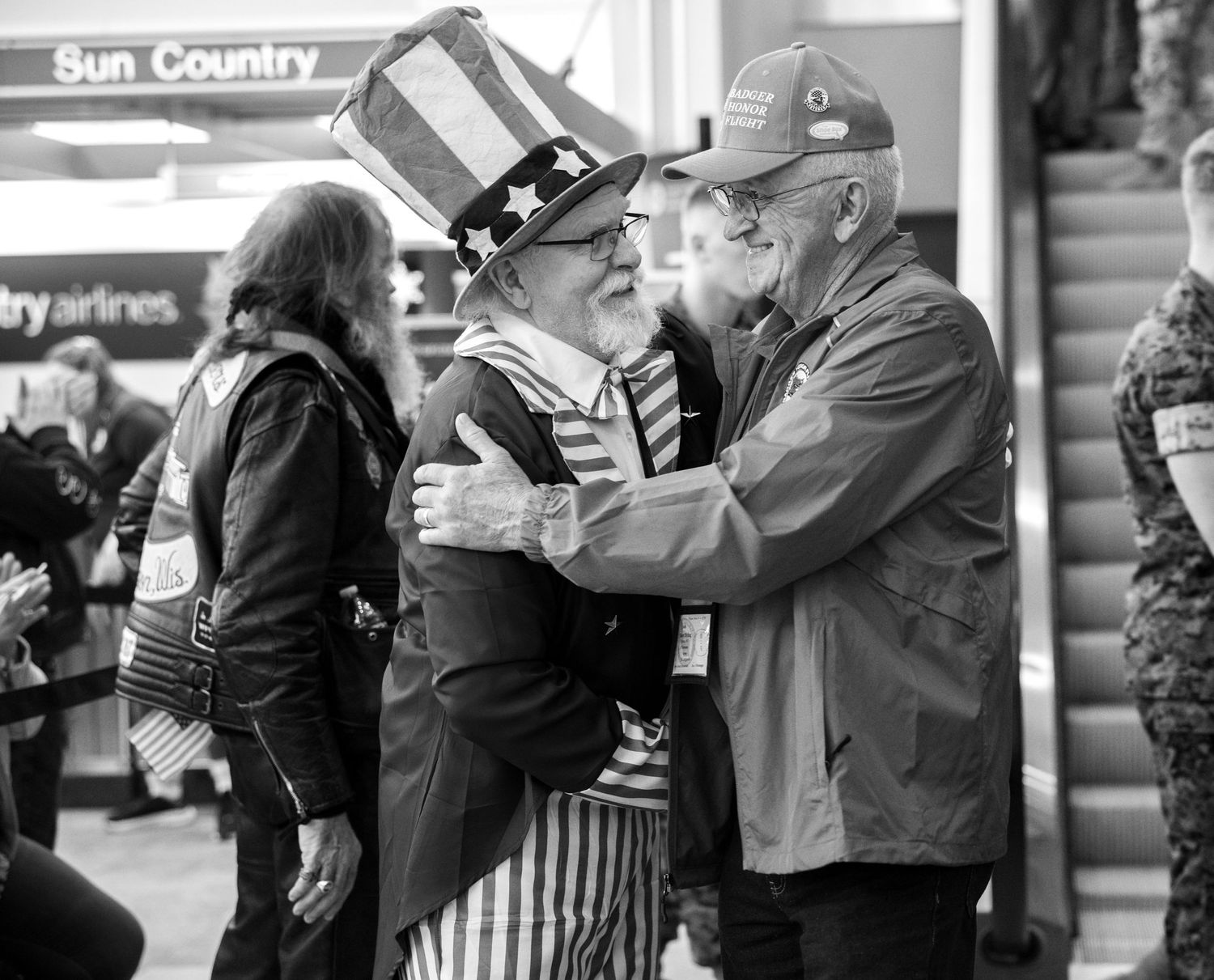 Two men hugging warmly at airport, one dressed as Uncle Sam in patriotic costume.