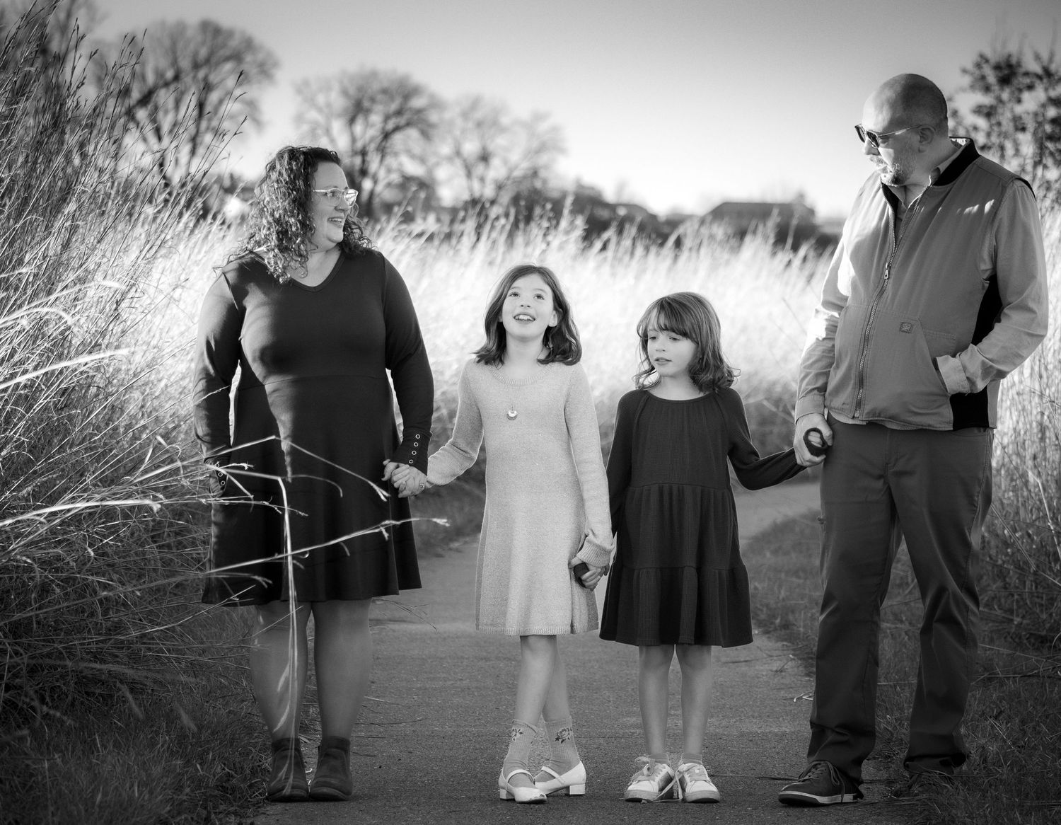 A happy family of four holds hands on a scenic outdoor path in a black and white portrait.