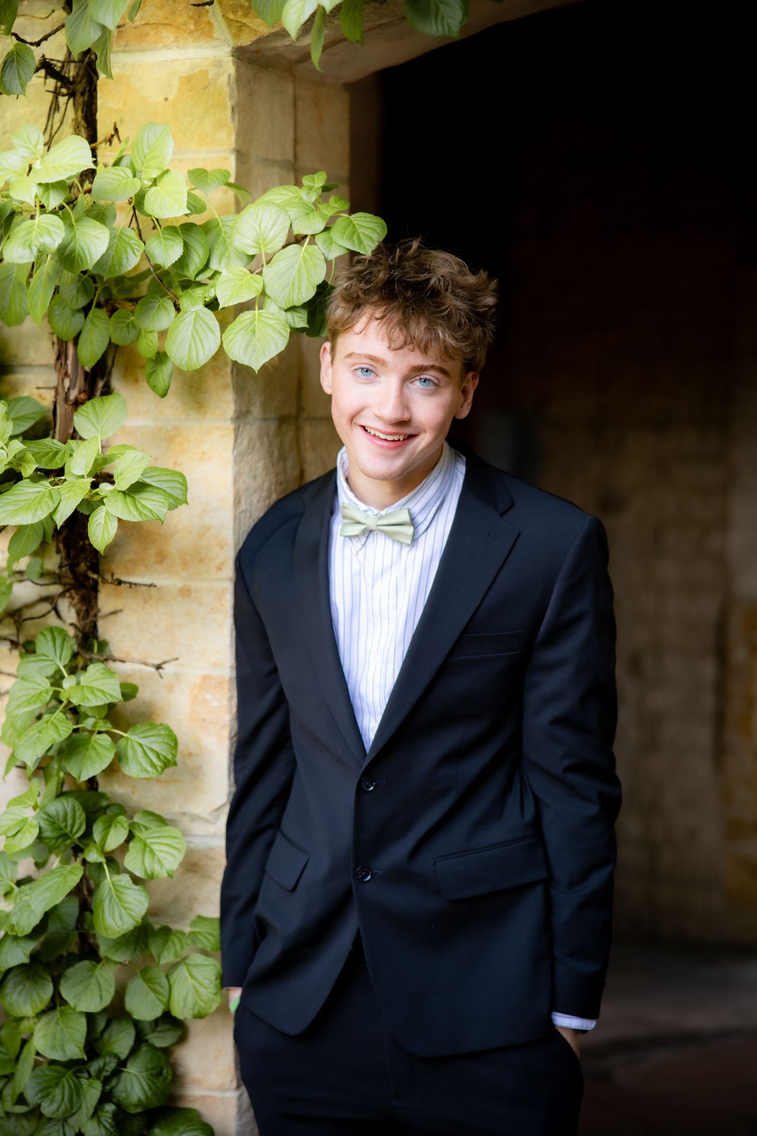 Smiling Senior in black suit and bow tie posing by stone wall with ivy.