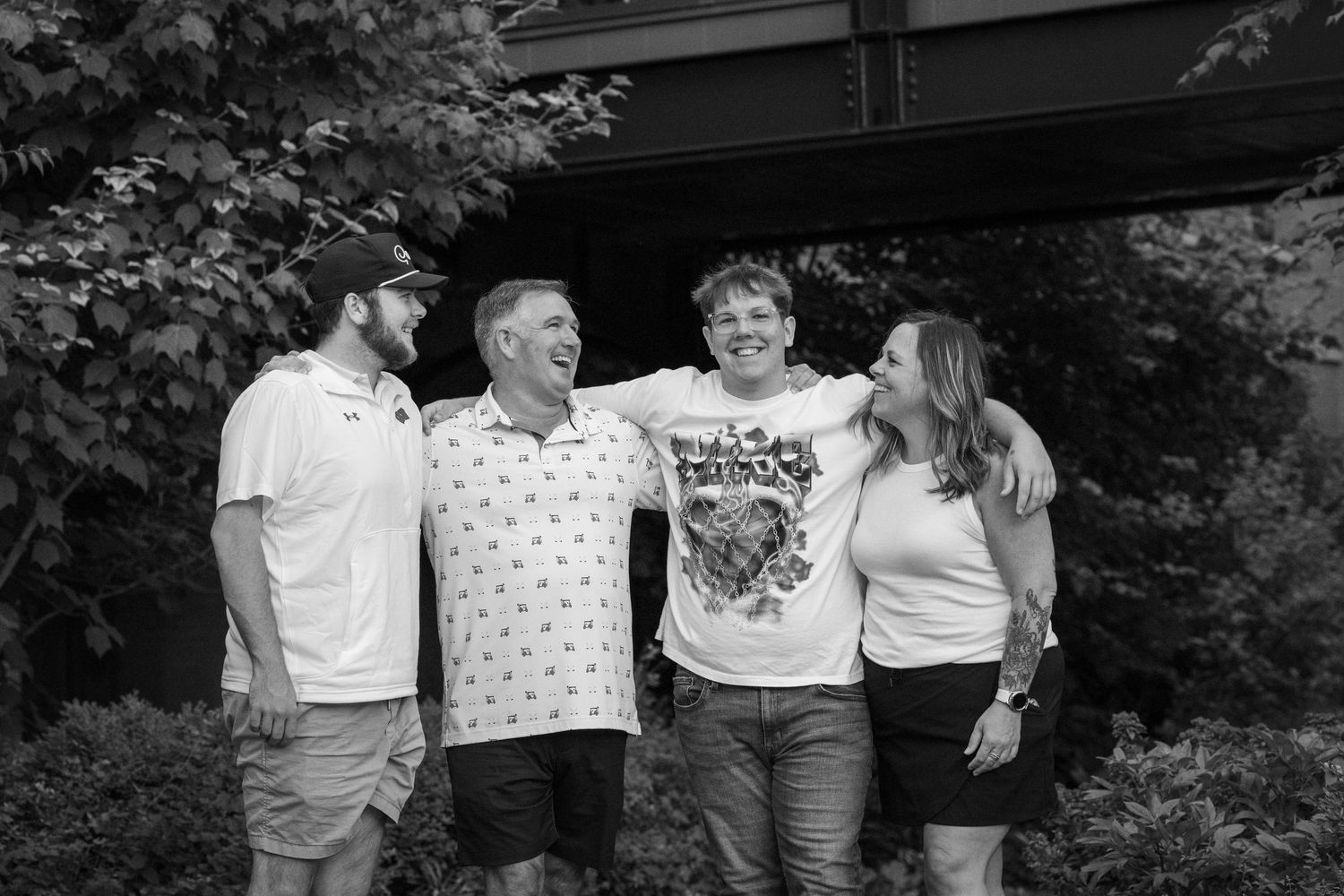 Black and white photo of four smiling adults posing together outdoors under a bridge surrounded by lush greenery.