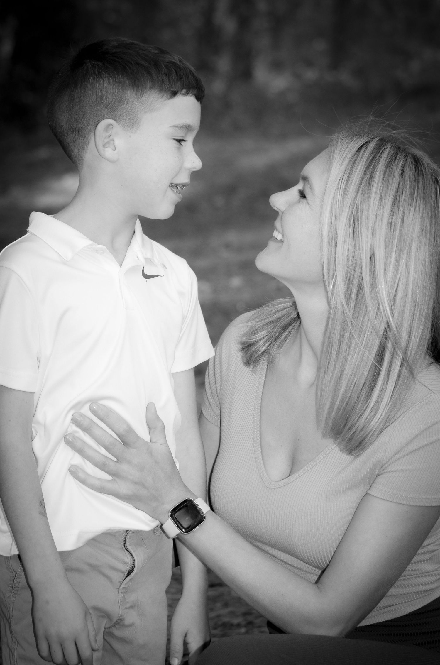 Black and white portrait of blonde mother smiling at her young son in white polo shirt outdoors.