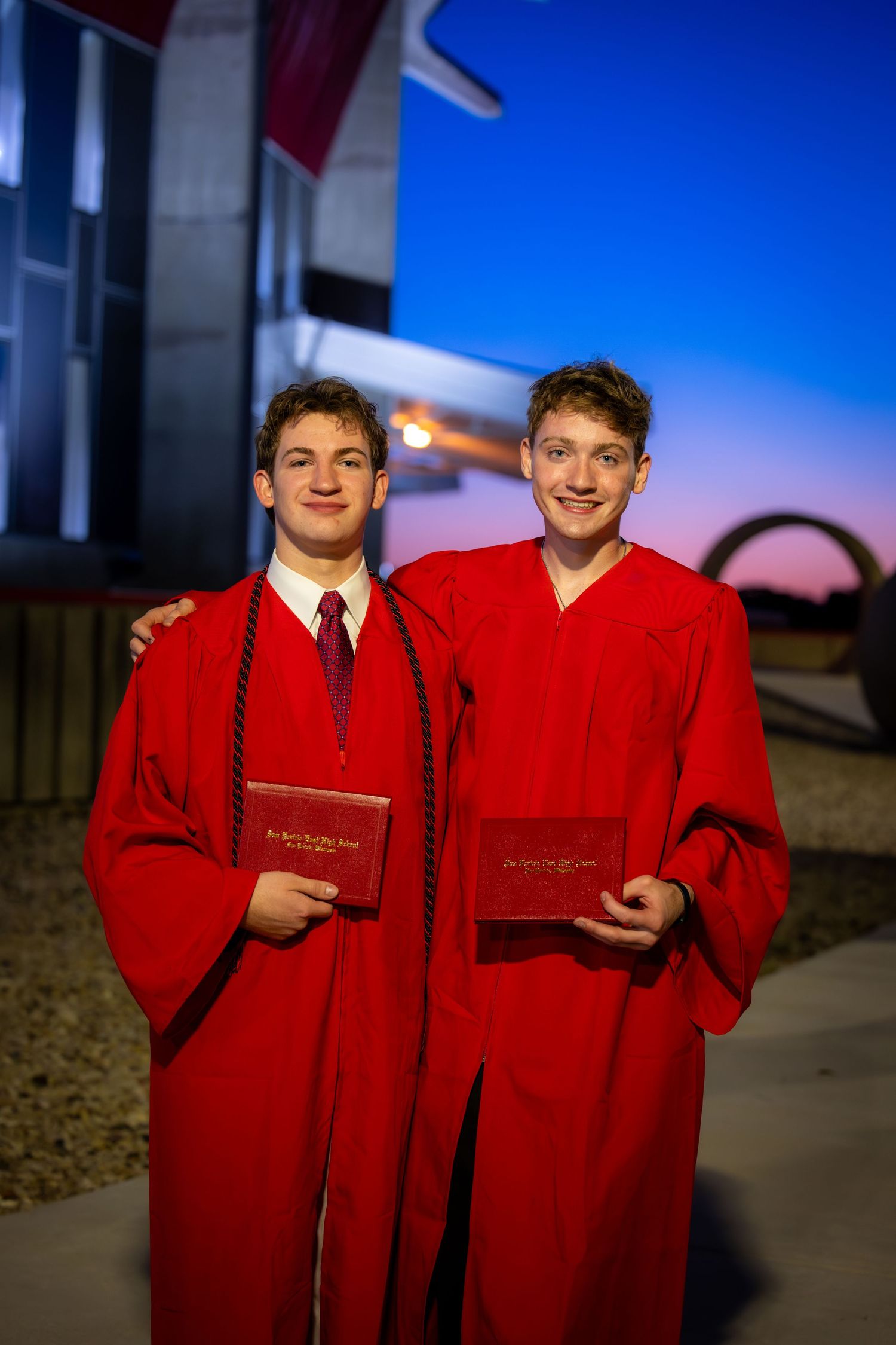 Two young men in red graduation gowns holding diplomas at sunset.