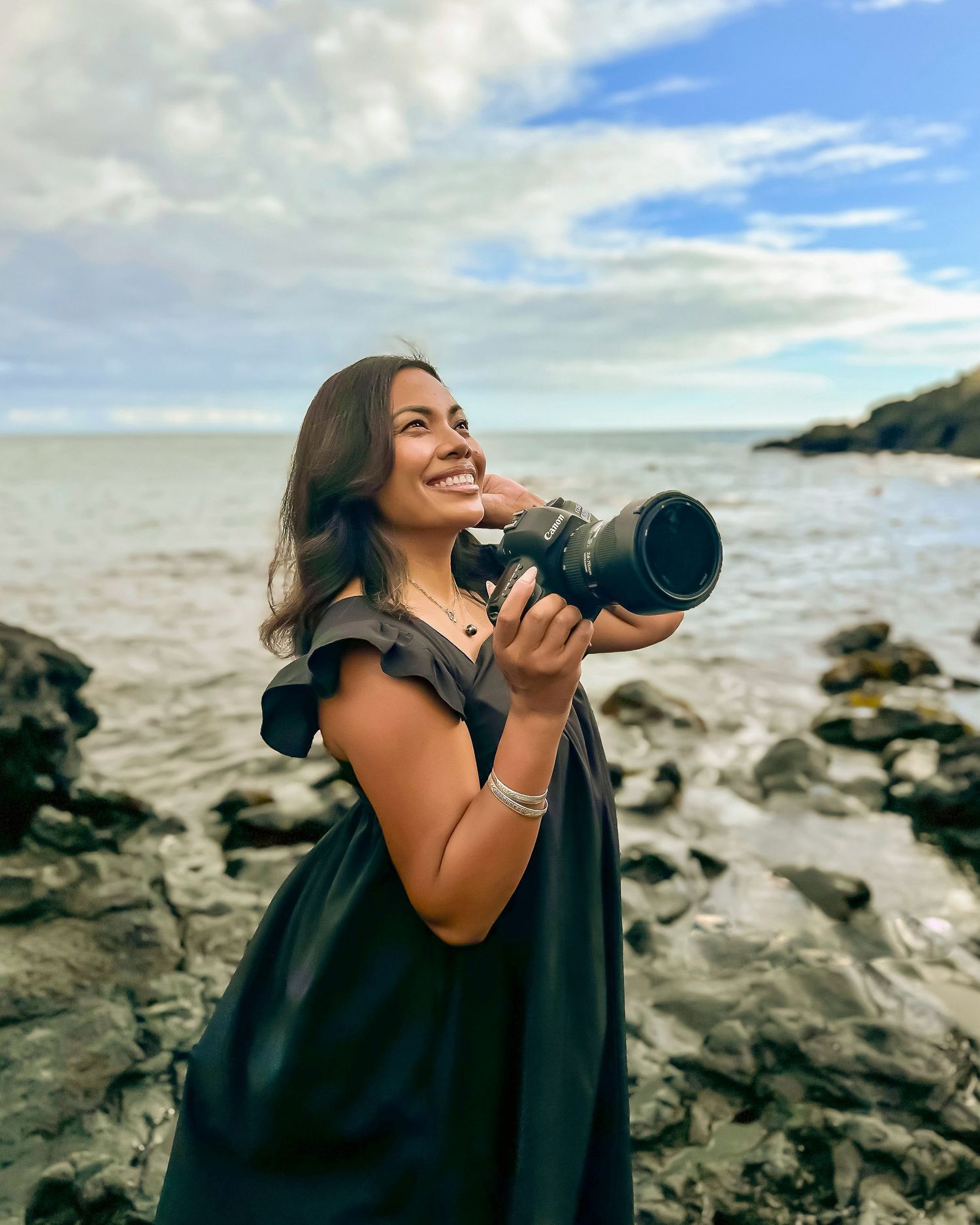 Photographer with camera capturing ocean views from rocky coastline under cloudy sky.