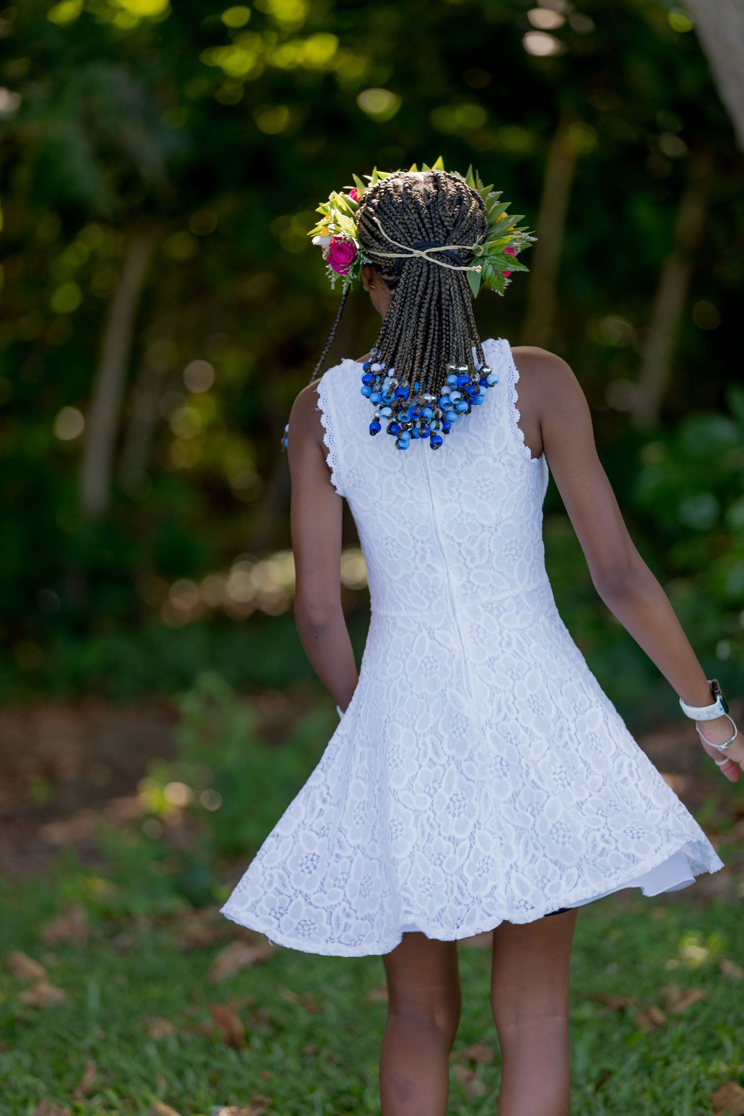 A person wearing a white lace sundress and floral crown spins in a garden setting with braided hair adorned with blue flowers.