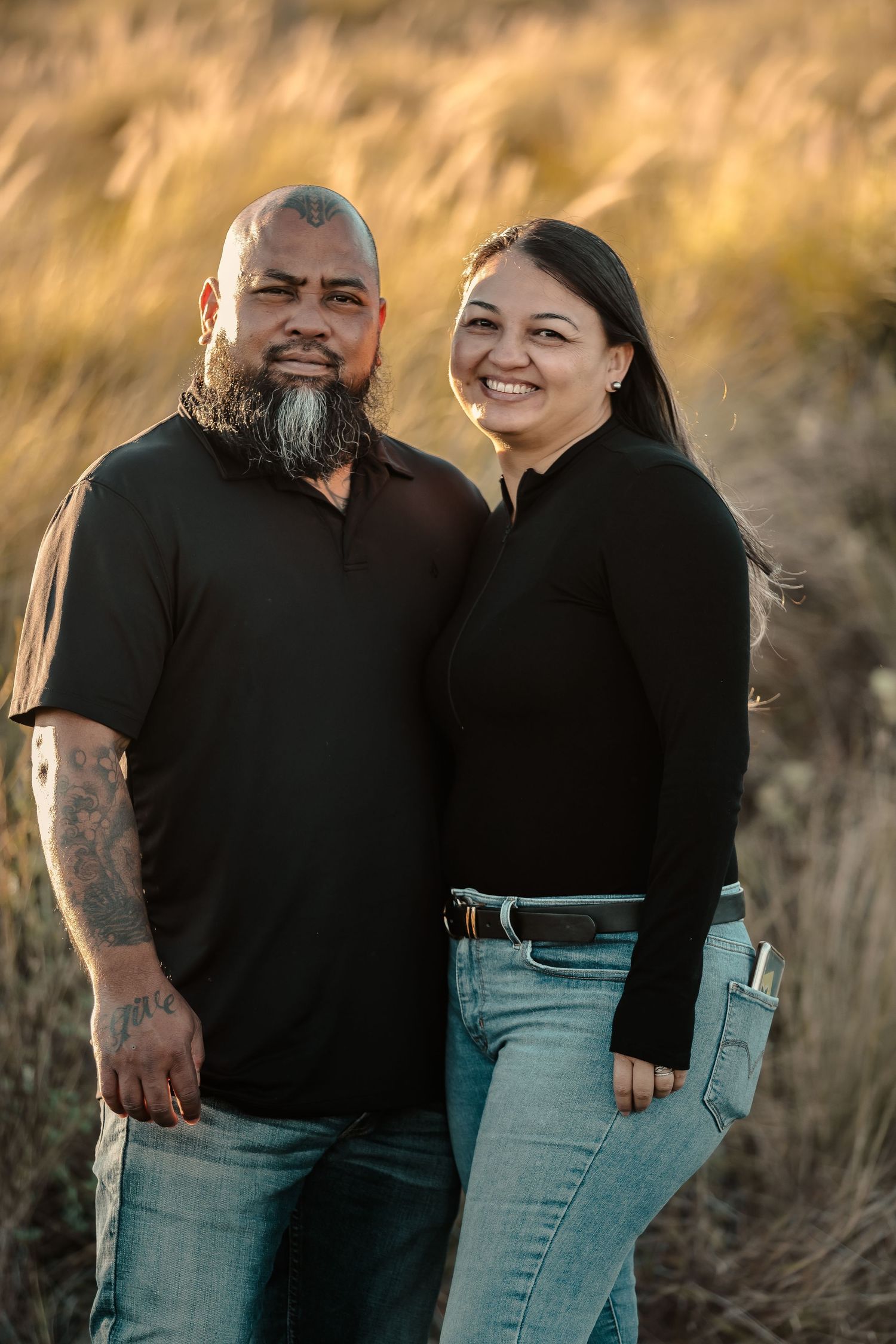 Couple poses together in casual attire against golden sunset field.