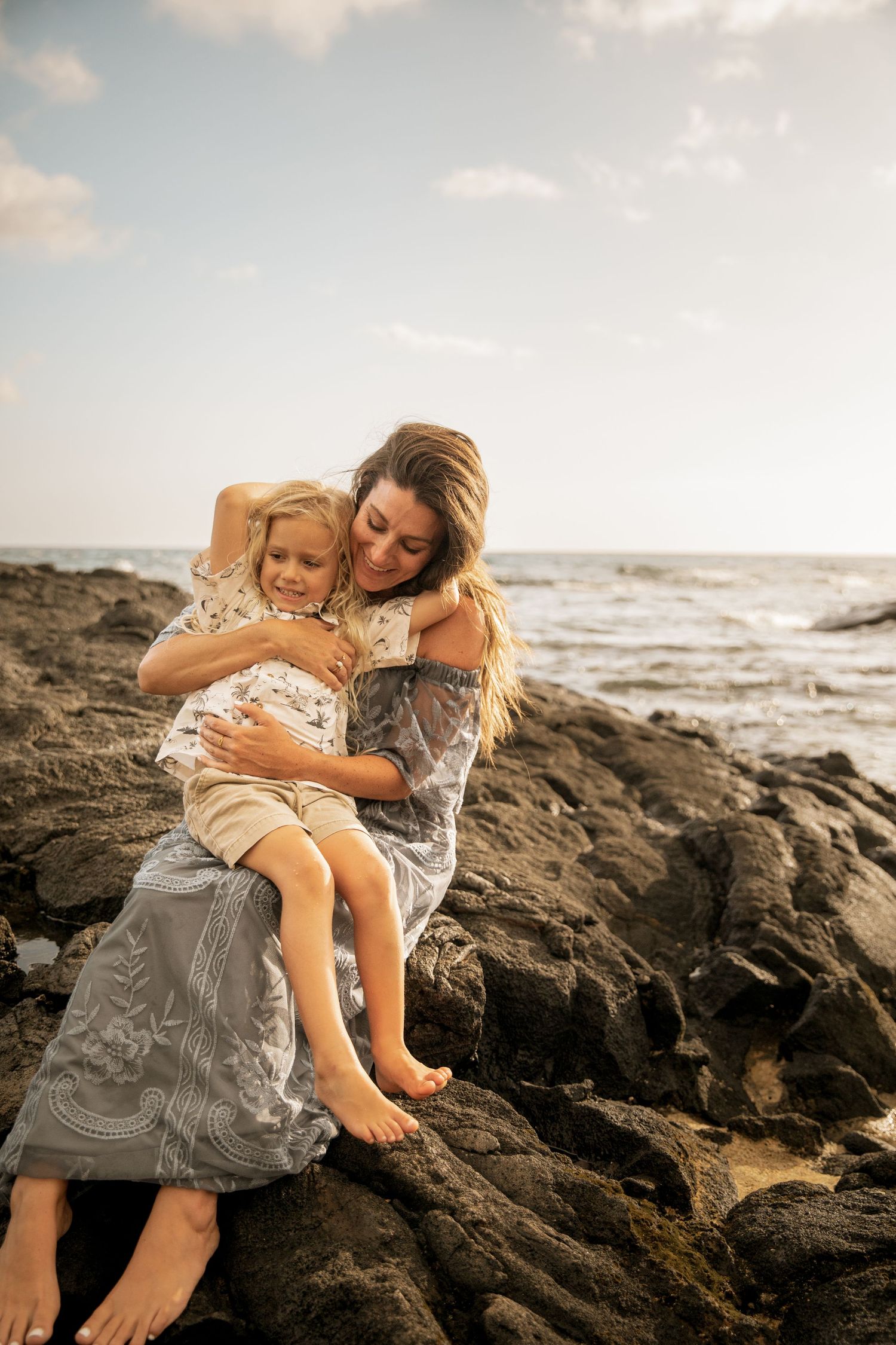 A tender moment shared between two people sitting on volcanic rocks by the ocean at sunset.