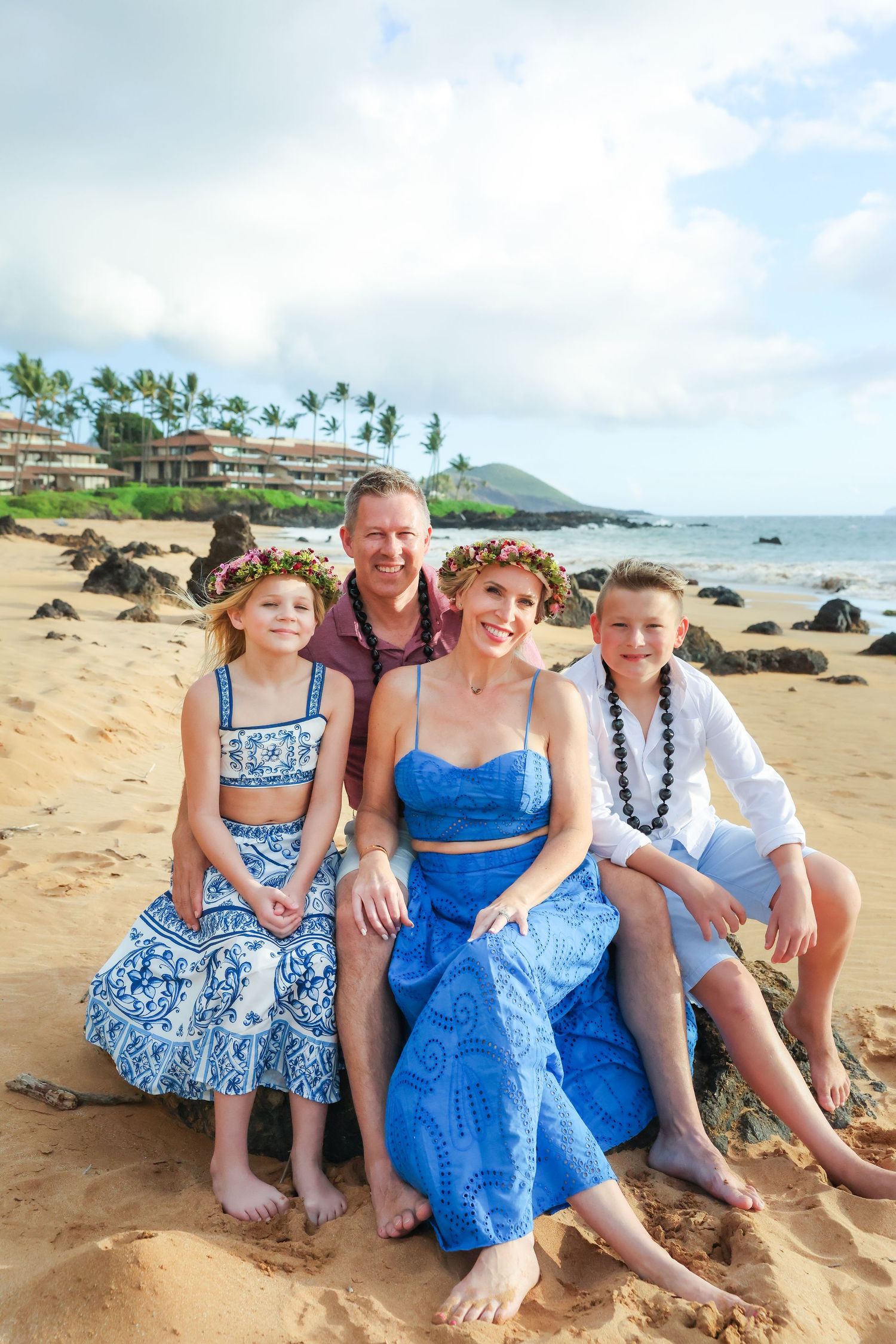 A family wearing coordinating blue outfits sits together on a sandy Hawaiian beach with ocean and palm trees in background.