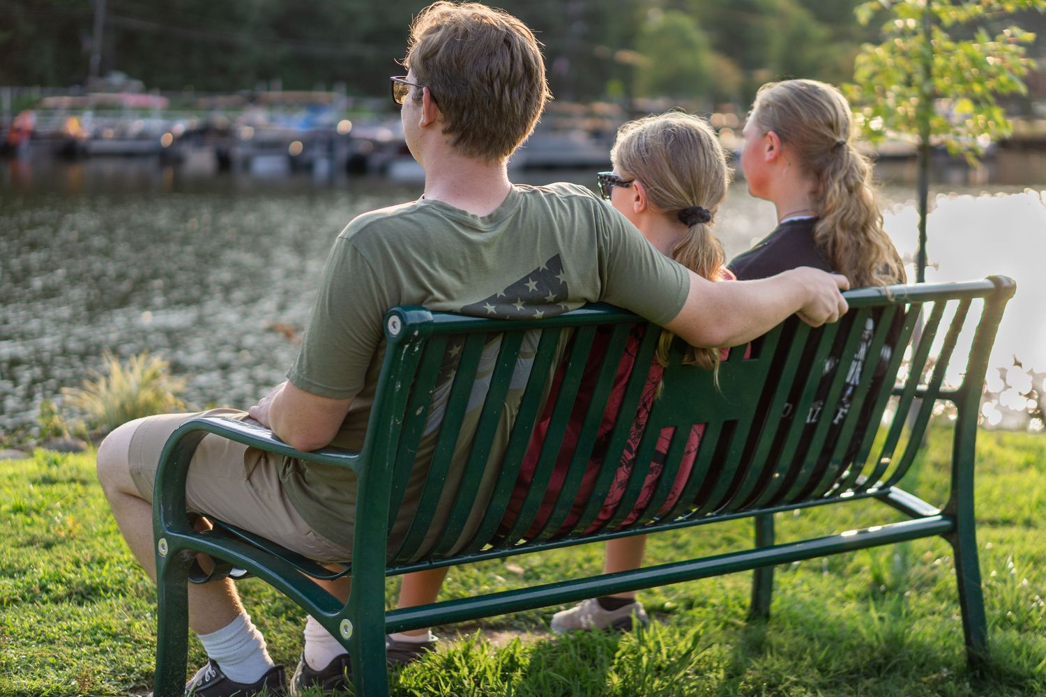 Three people sitting on a green park bench overlooking a calm lake surrounded by lush greenery.