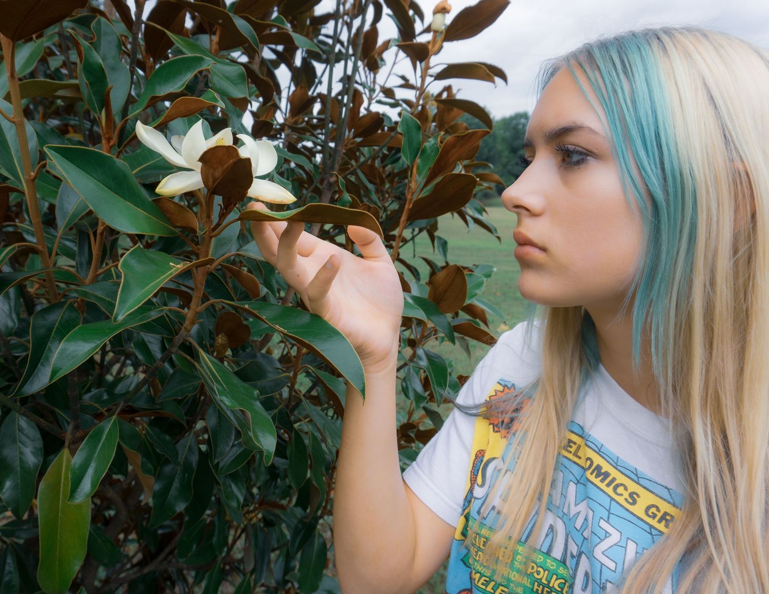 Young woman with blue-tipped blonde hair examining a white magnolia flower on a green leafy shrub.
