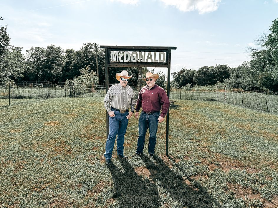 Two men standing at the entrance of McDonald Ranch under a rustic sign, surrounded by green fields and trees.