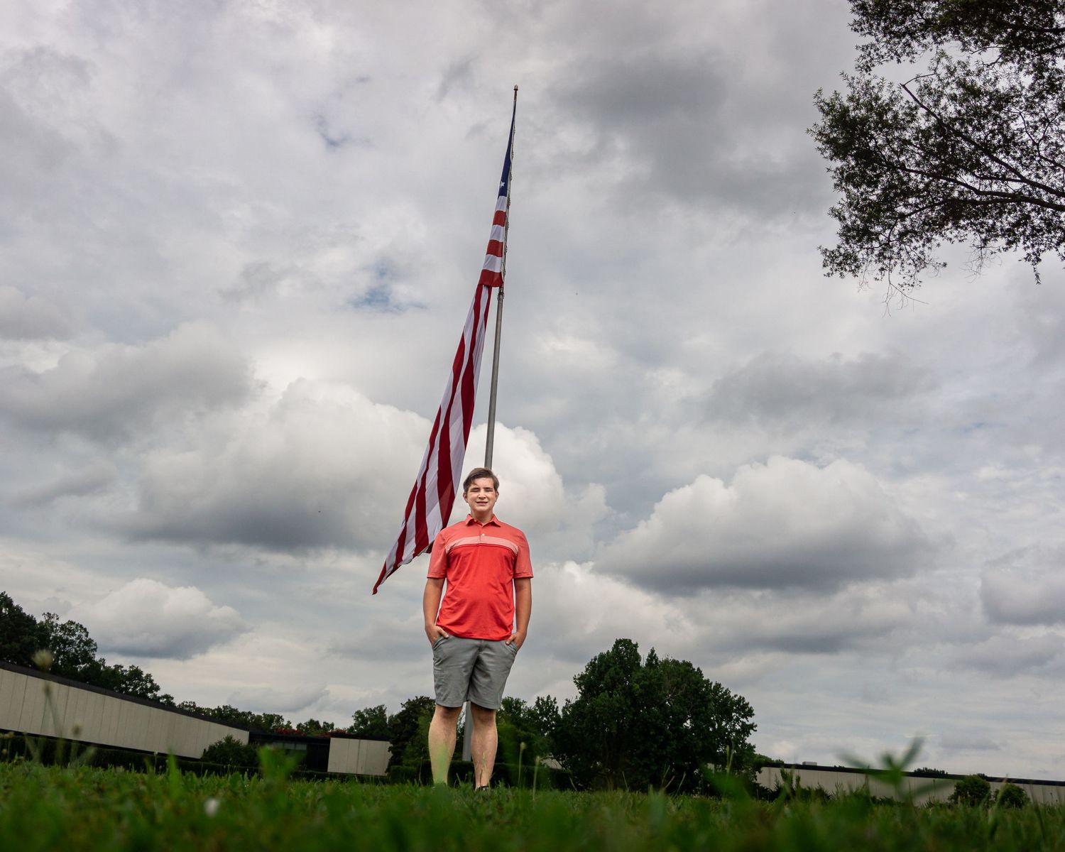 Young boy in red shirt stands proudly before a tall American flag on green grass under cloudy skies.
