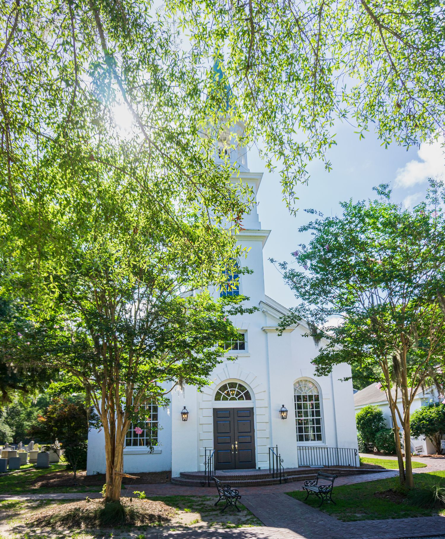 White colonial church with steeple surrounded by lush green trees on a sunny day.