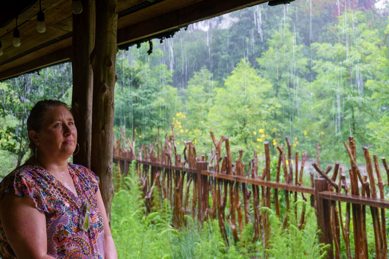 Woman standing on a rustic wooden porch watching rain fall over a lush green forest.