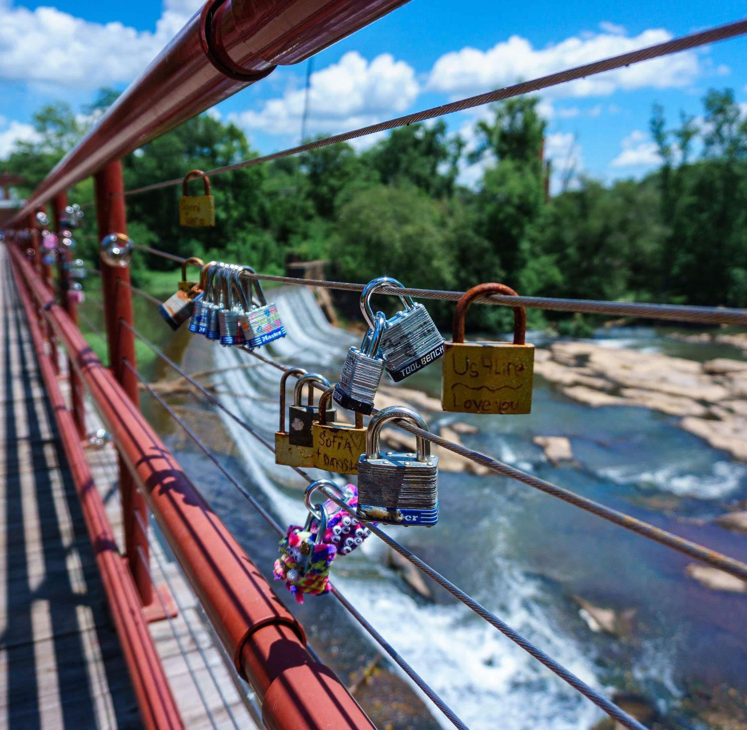 Love locks attached to a red suspension bridge railing over a rocky river, surrounded by lush green trees under a blue sky.
