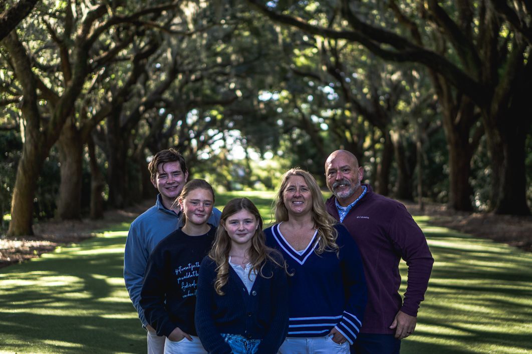 Family of five poses together on a grassy path lined with large oak trees draped in dappled sunlight.