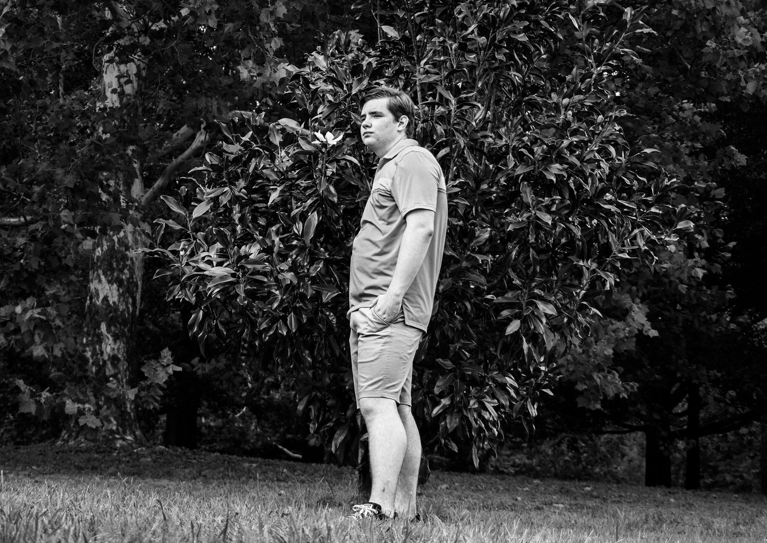 Black and white photo of a young man standing on grass in front of lush green trees outdoors.