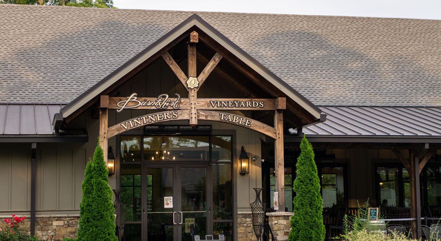Rustic winery entrance with timber frame architecture, stone accents, and manicured evergreen trees flanking glass doors.