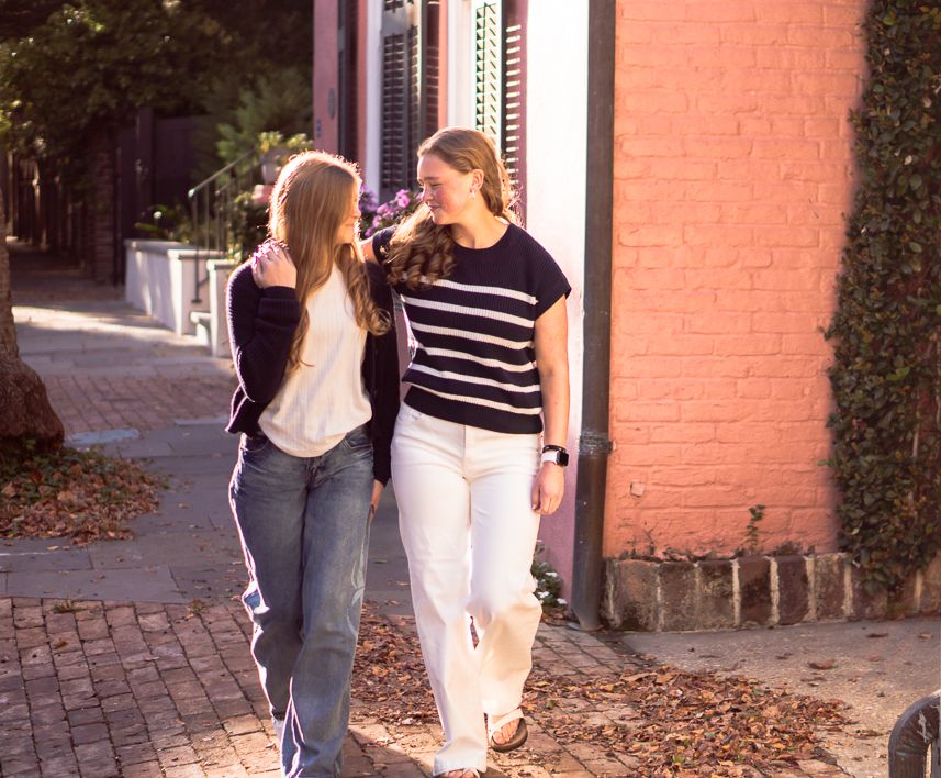 Two young women walk and talk along a brick sidewalk lined with fallen leaves, one in a striped top and white pants, the other in jeans.