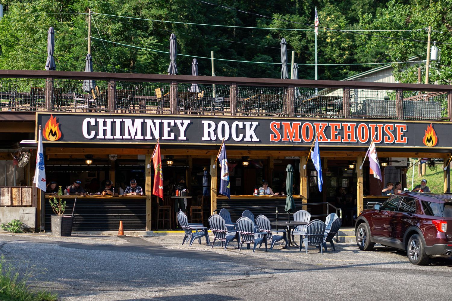 Chimney Rock Smokehouse restaurant exterior with outdoor seating, flame signage, and lush green trees in background.