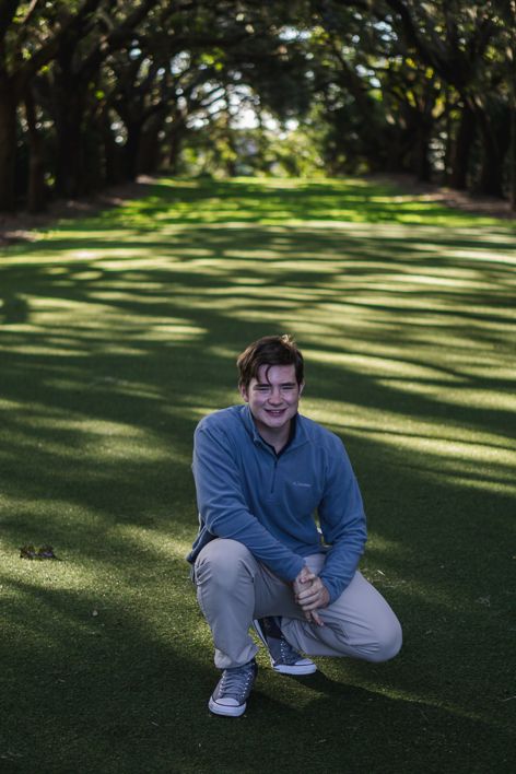 Smiling young man in a blue pullover and khaki pants crouches on a sunlit grass lawn beneath an alley of shady oak trees.