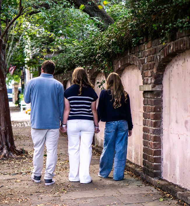 Three people walk away from the camera along a tree-lined sidewalk beside a weathered pink brick wall with arched details.