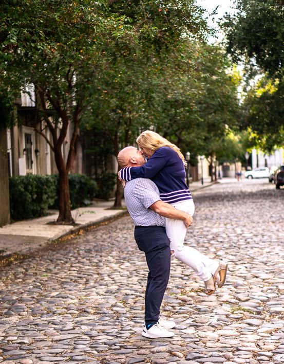 A man lifts a smiling woman in an embrace on a cobblestone street lined with leafy green trees and historic buildings.