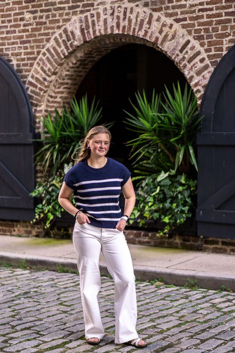 Young woman in a navy striped short-sleeve top and white wide-leg pants standing on a cobblestone street before a brick archway with green p