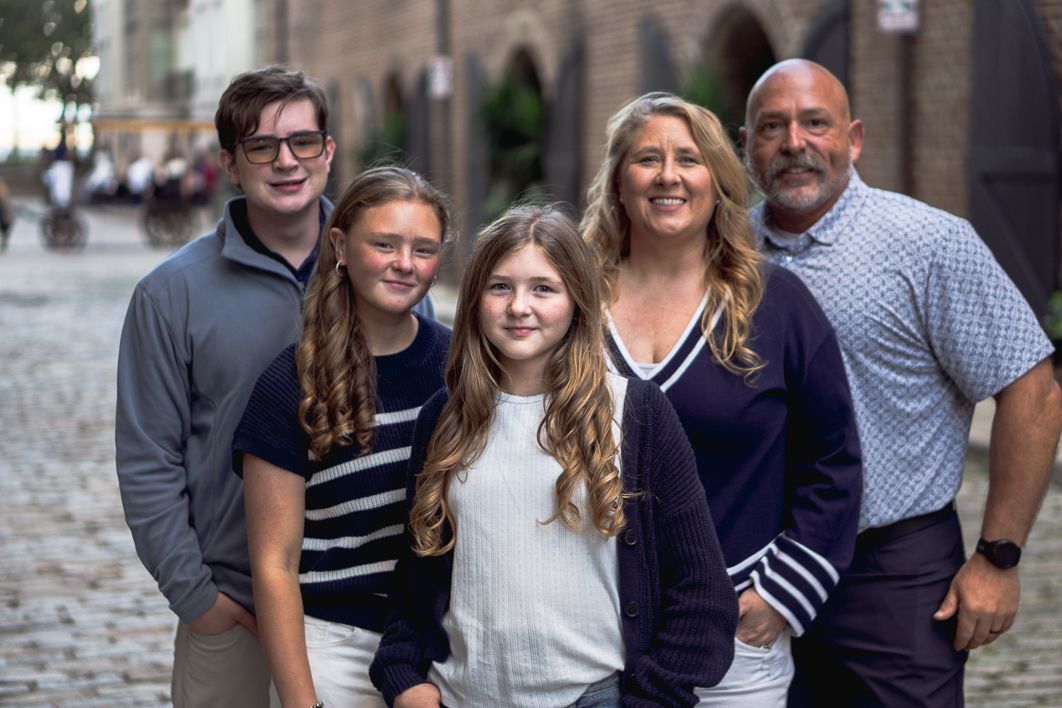 Family of four smiling together outdoors on a cobblestone street, with two teens, a woman and a man posing in casual coordinated clothing.