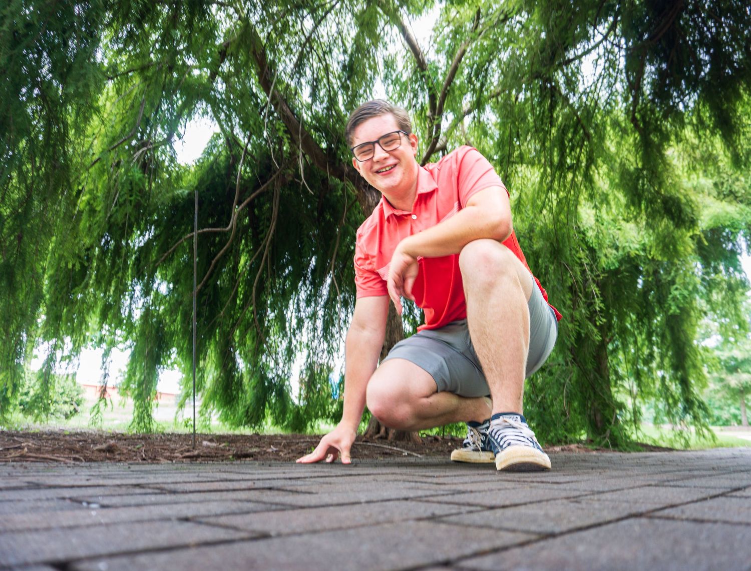 Young man in red polo shirt crouching under weeping willow tree on brick path, smiling at camera outdoors.