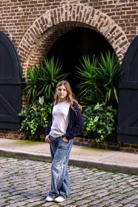 Young girl in a white top, navy cardigan and wide-leg jeans poses on a cobblestone street before a brick archway with green plants.