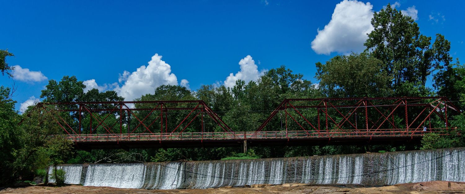 Historic red iron truss bridge spans a concrete dam waterfall surrounded by lush green trees under a vivid blue sky.
