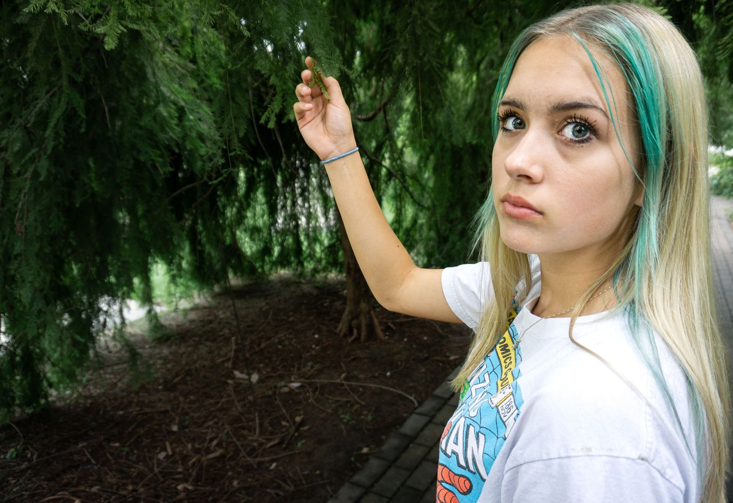 Young woman with blonde and teal hair holding tree branch outdoors in lush green park setting.